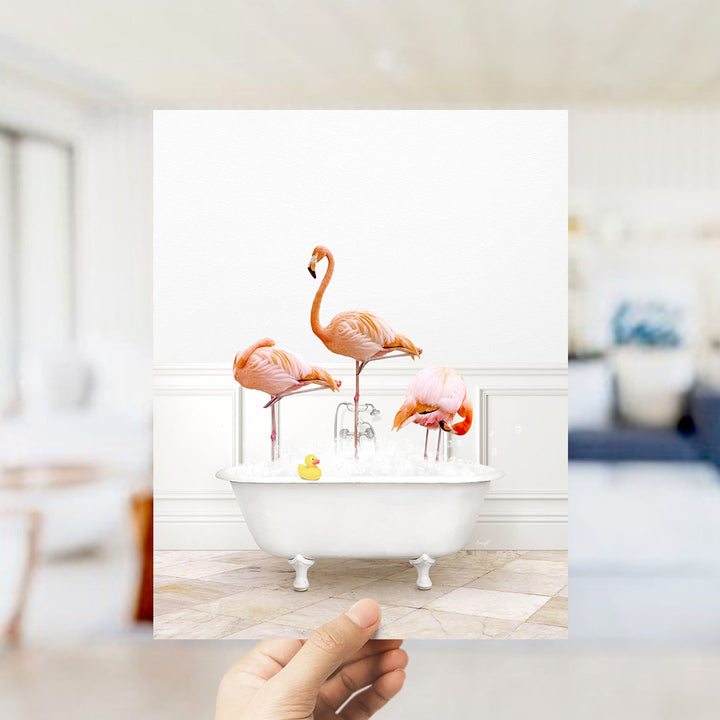 A hand holds up a framed image depicting three flamingos standing in a white bathtub filled with water and floating on their hind legs.
