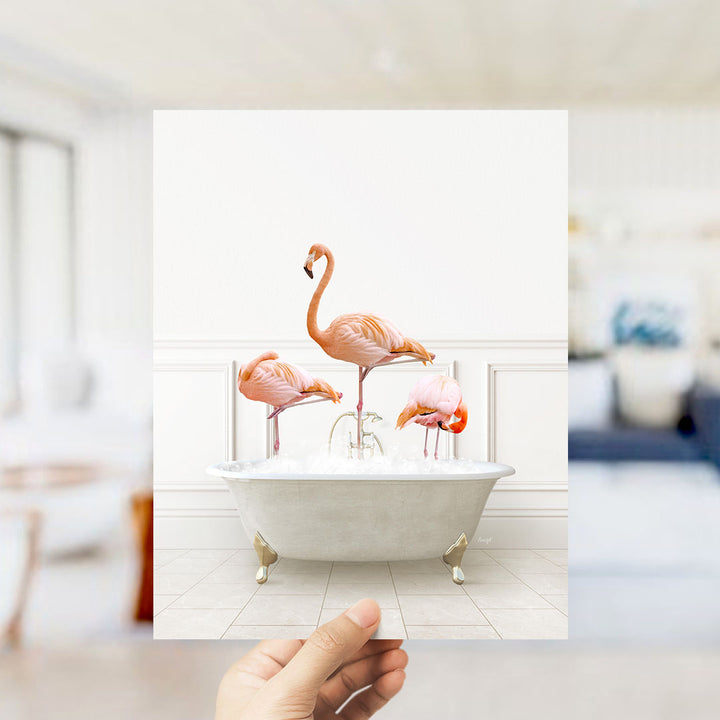 A hand holds up a framed photograph of three flamingos standing in a white bathtub filled with water.