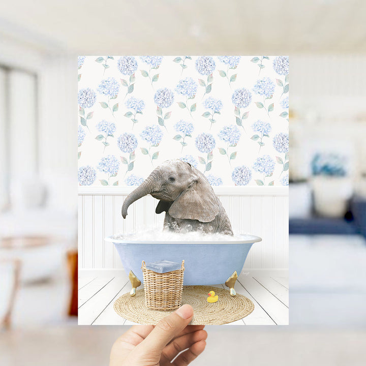 A hand holds up a framed image depicting a baby elephant sitting in a blue bathtub filled with water, surrounded by a blue and white floral pattern.