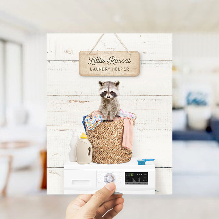 A hand holding a sign that says "Little raccal Laundry helper" with a raccoon sitting in a laundry basket.