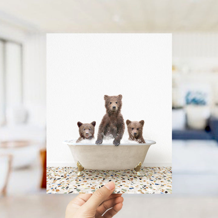 A hand holds up a framed photograph of three adorable brown bears sitting in a bathtub filled with water.