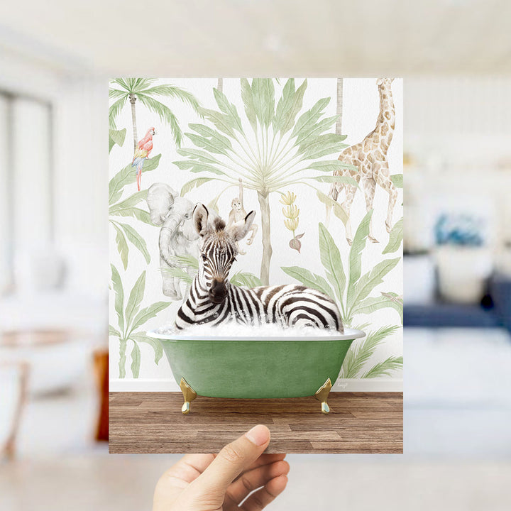 A hand holds up a framed print featuring a zebra relaxing in a green bathtub surrounded by tropical foliage, including a giraffe and palm trees.