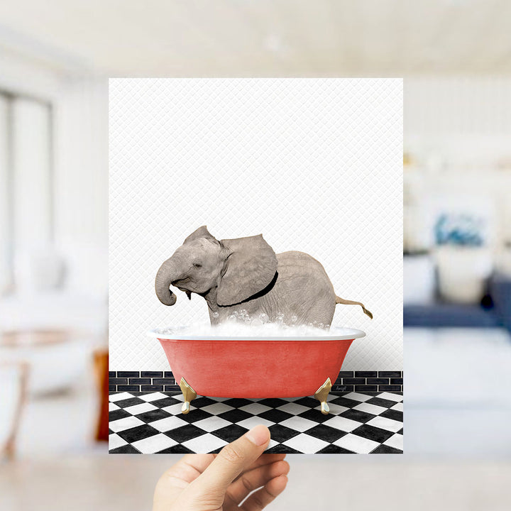 A hand holds up a framed image depicting a gray elephant sitting in a red bathtub filled with bubbles.