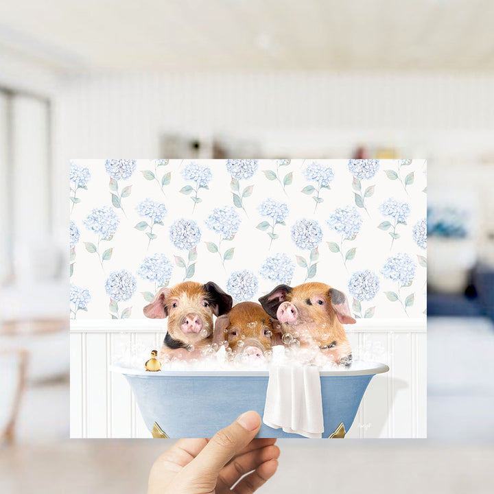 A hand holding a framed image of three adorable pigs sitting in a bathtub filled with bubbles, surrounded by a floral pattern.