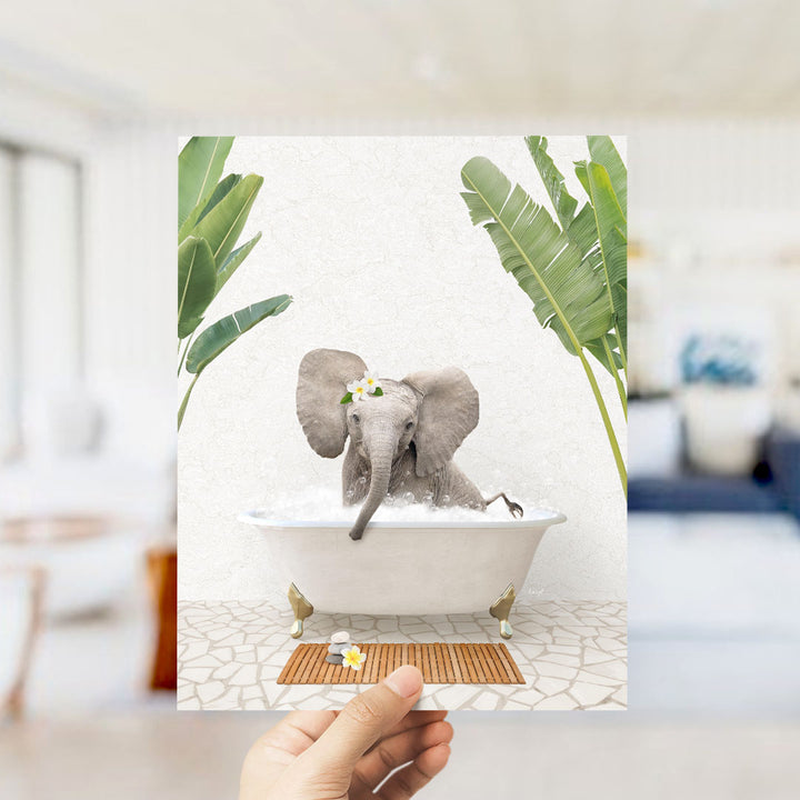 A hand holds up a framed image depicting a baby elephant sitting in a bathtub, surrounded by tropical plants.