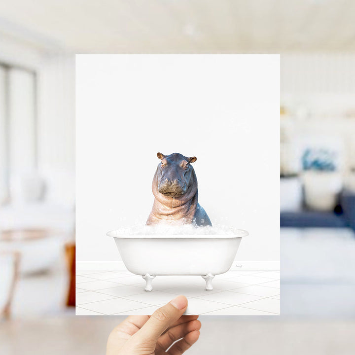 A hand holds up a framed image of a hippopotamus sitting in a bathtub filled with water, creating a whimsical and playful scene.