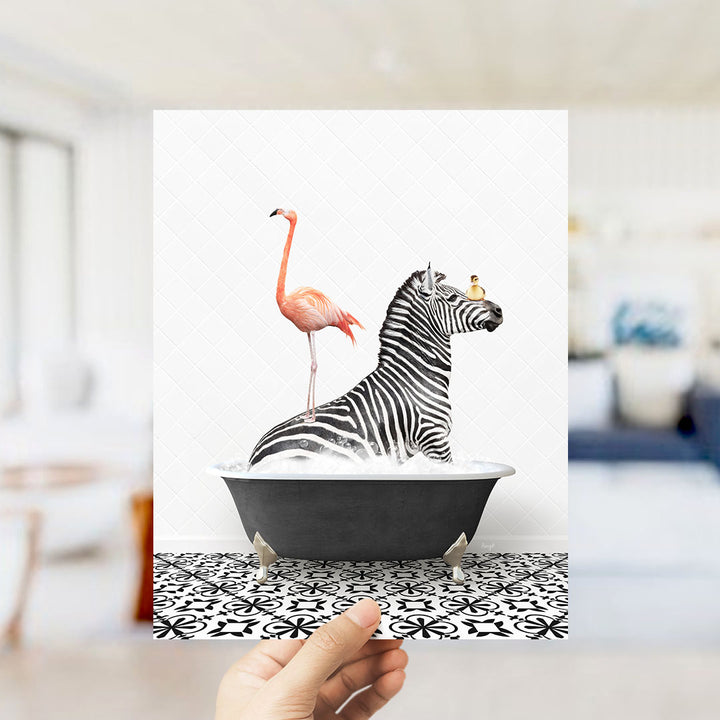 A hand holds up a framed image depicting a zebra standing on one leg inside a bathtub, with a pink flamingo perched on its back.