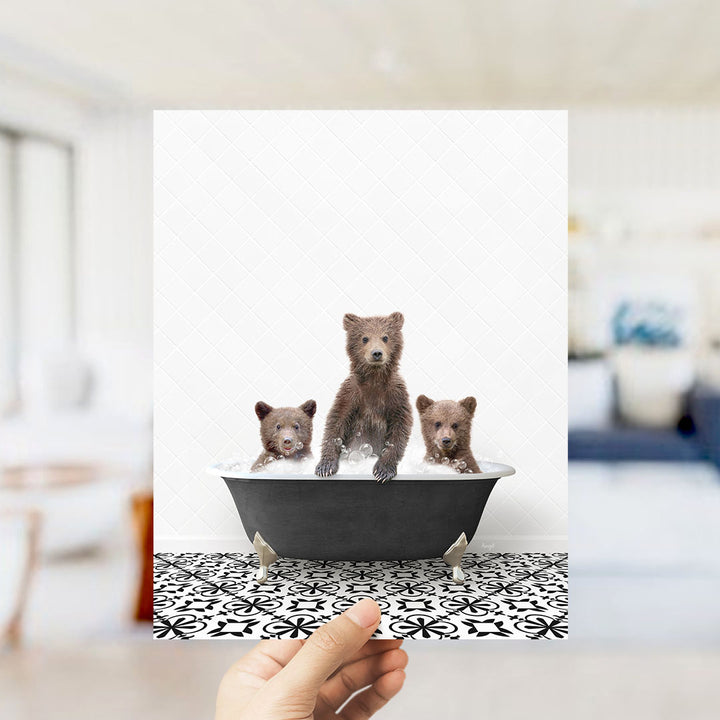 A hand holds up a framed image of three adorable brown bears sitting in a bathtub, with one bear sitting in the center and the other two on either side.