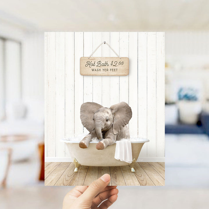 A hand holding a framed image of a baby elephant sitting in a bathtub, with a sign above it that says "Hot Bath $2.00 WASH YOUR FEET".