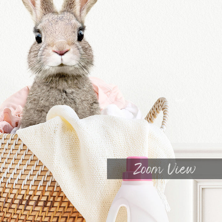 A cute gray rabbit peeking out from a woven basket filled with laundry, including a white towel and a plastic bottle.