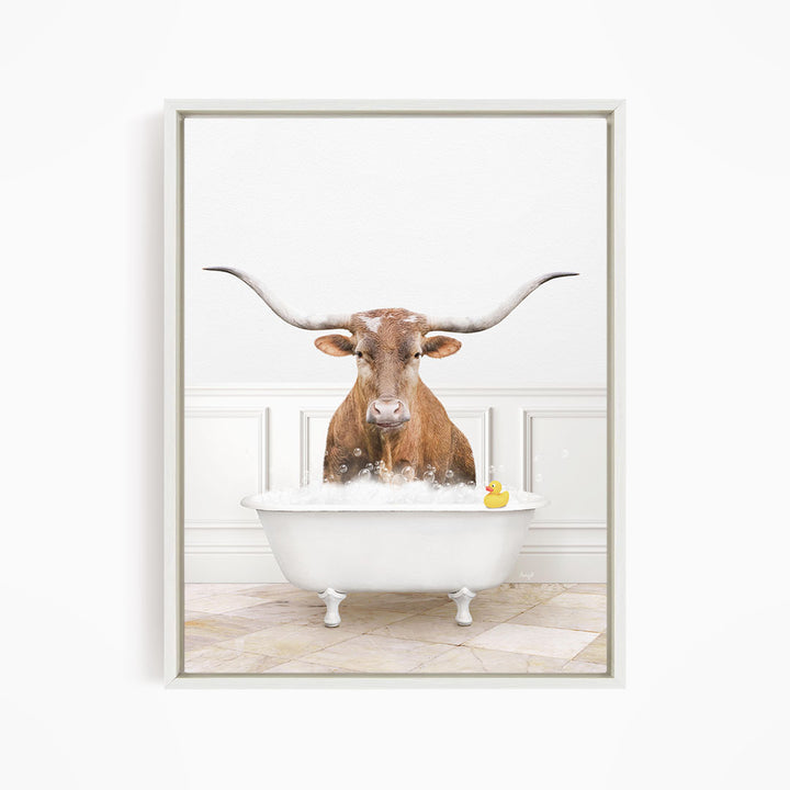 A brown cow with long horns is sitting in a white bathtub filled with water, with a yellow rubber duck floating in the water.
