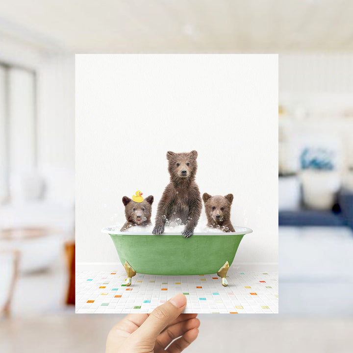 A hand holds up a framed image depicting three adorable brown bears sitting in a green bathtub filled with water, surrounded by colorful tiles on the floor.