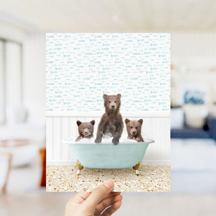 A hand holds up a collage of three images featuring three adorable brown bears sitting in a bathtub filled with water, surrounded by a fish-themed wallpaper.