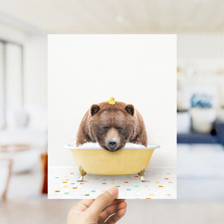 A hand holds up a framed image of a brown bear sitting in a yellow bathtub, with a rubber duck floating on its head.