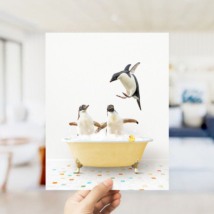 A hand holds up a framed image depicting two penguins sitting in a yellow bathtub filled with bubbles, with one penguin standing on its hind legs and another sitting on its belly.