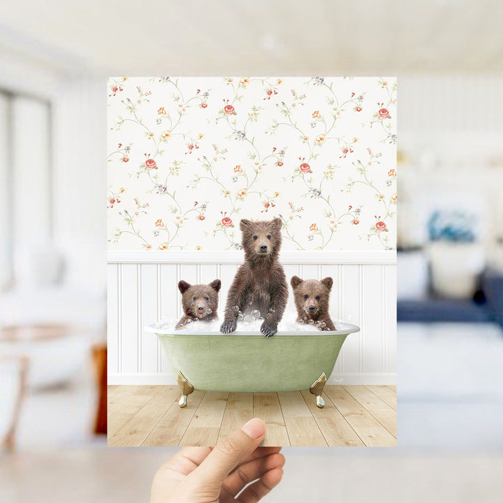 A hand holds up a framed photograph of three adorable brown bears sitting in a green bathtub, with a floral wallpaper in the background.
