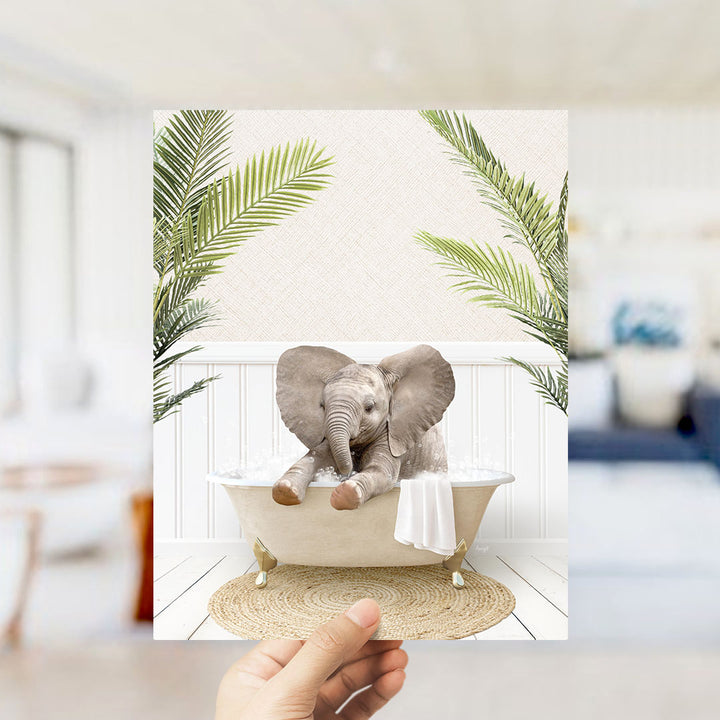 A hand holds up a framed image depicting a baby elephant sitting in a bathtub, surrounded by palm leaves.