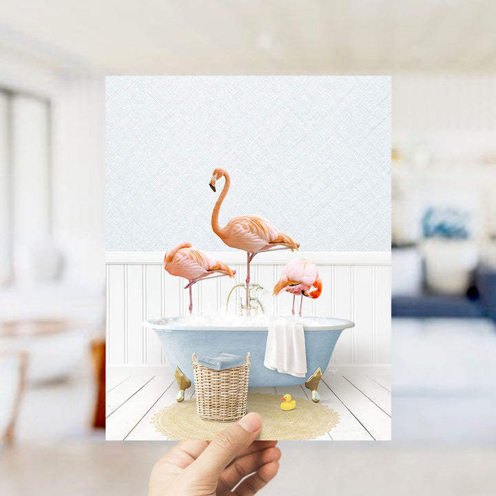 A hand holds up a framed image depicting three flamingos standing in a bathtub filled with bubbles, surrounded by a basket of bath toys.