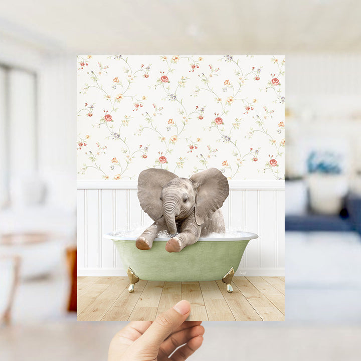 A hand holds up a framed image of a baby elephant sitting in a green bathtub, with a floral wallpaper in the background.