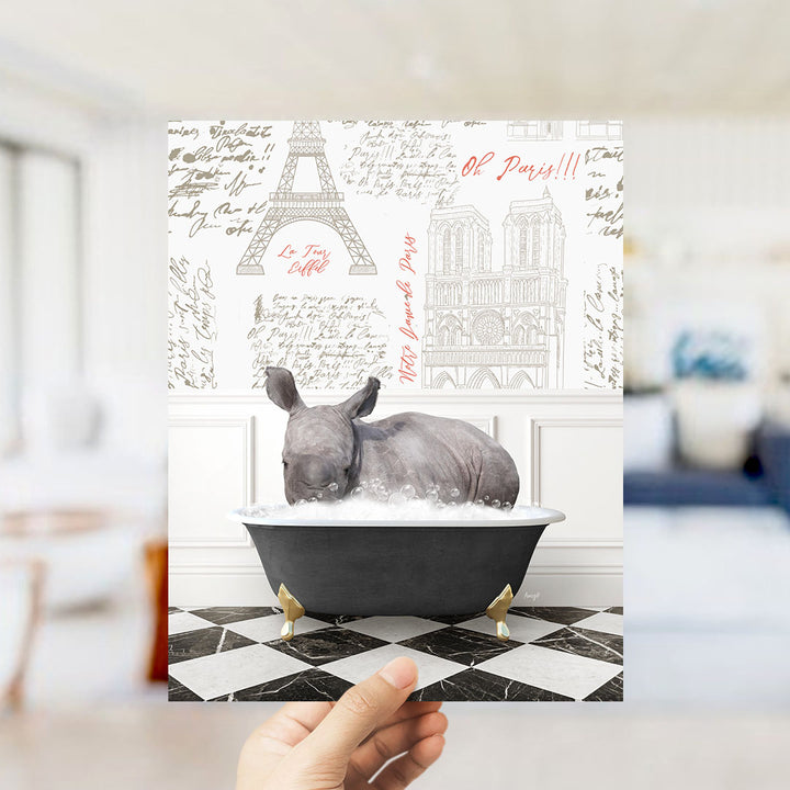 A hand is holding up a printed image featuring a rhinoceros sitting in a bathtub, surrounded by various Paris-themed elements such as the Eiffel Tower and the Notre-Dame Cathedral.