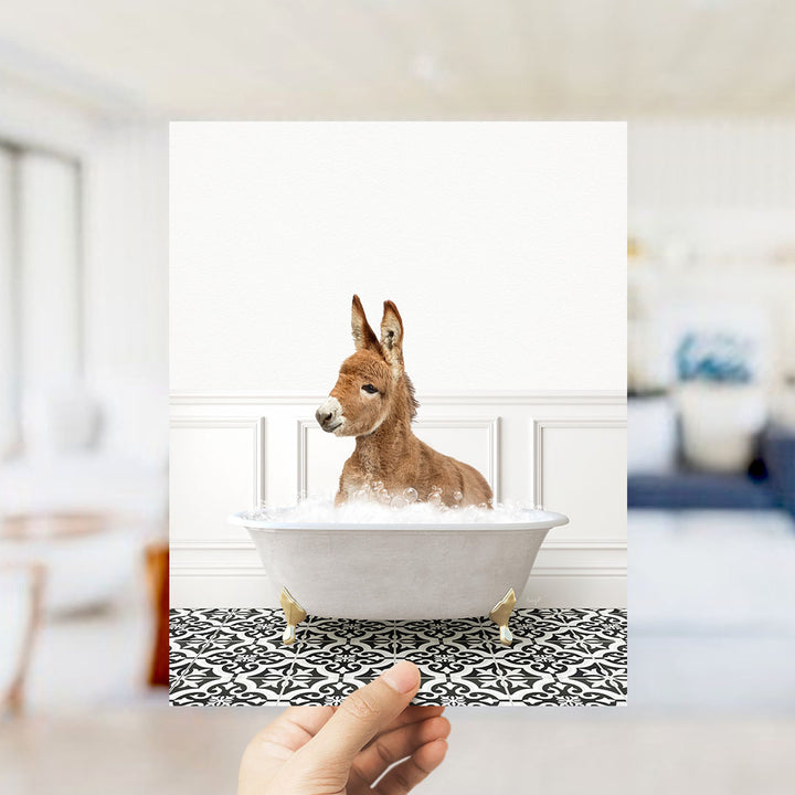 A donkey sitting in a white bathtub filled with bubbles, with a person's hand holding the photo in the foreground.