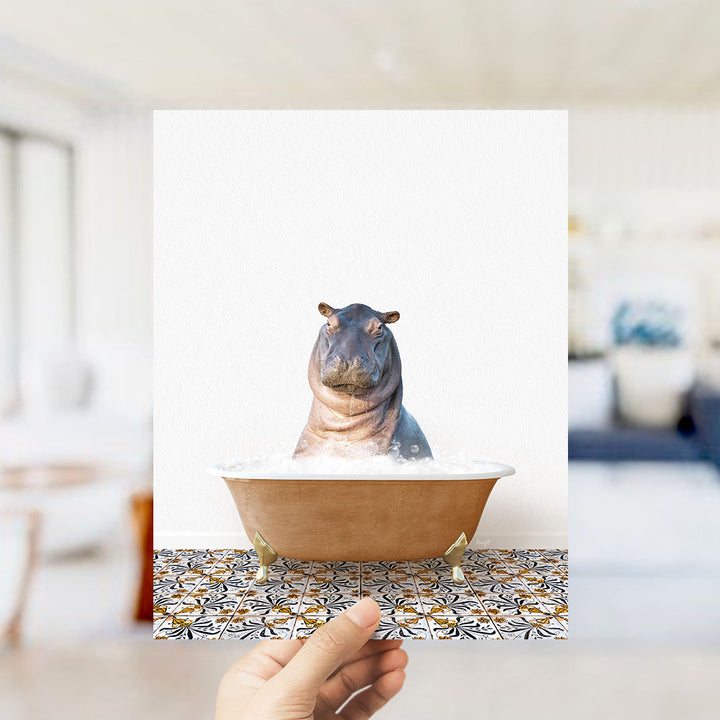 A hand holds up a framed image of a hippopotamus sitting in a bathtub, surrounded by a patterned tile floor.