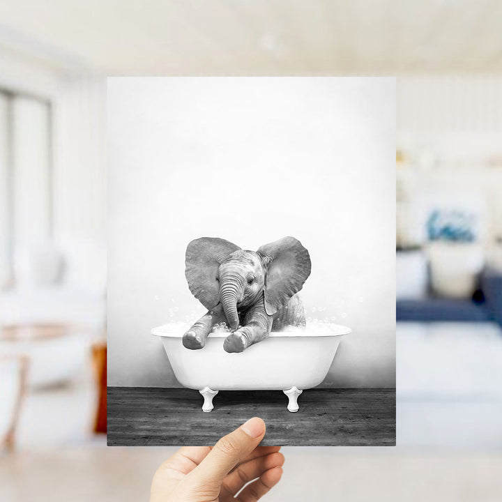 A hand holds up a black and white photograph of a baby elephant sitting in a bathtub, surrounded by a blurred background.