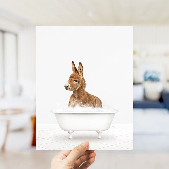 A hand holds up a framed image of a donkey sitting in a bathtub filled with bubbles.