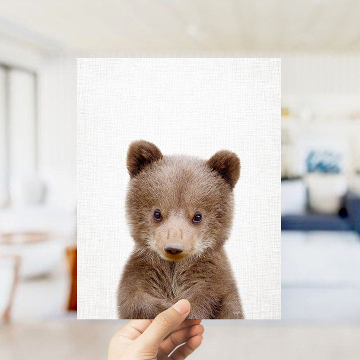 A hand is holding up a square print of a cute brown bear with a white background.