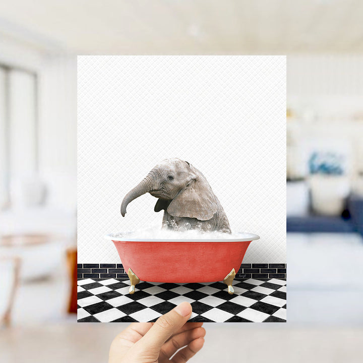 A hand holds up a framed image depicting a baby elephant sitting in a red bathtub filled with water, surrounded by a black and white checkered floor.