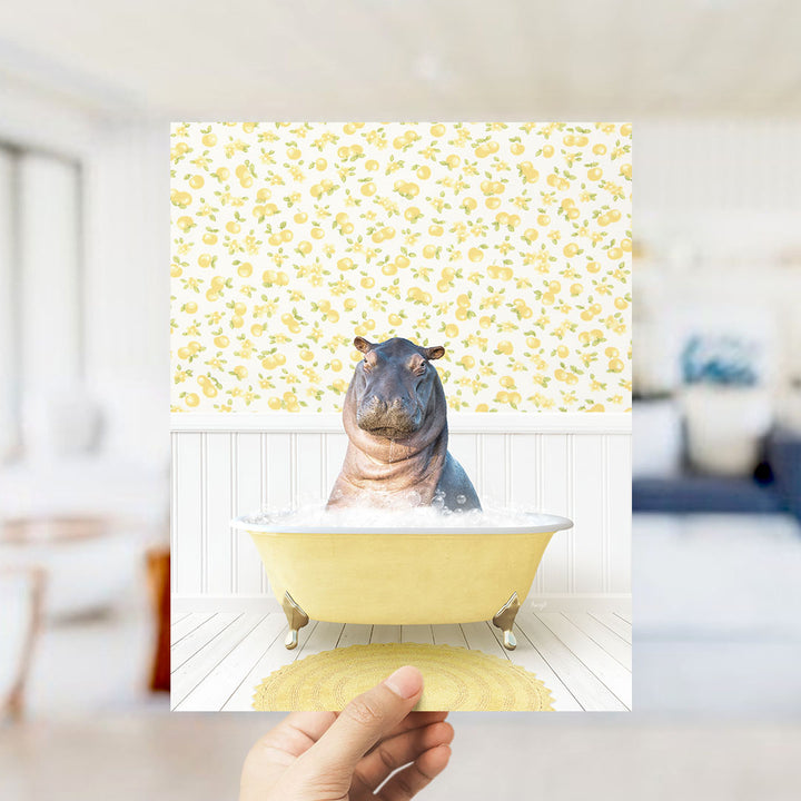 A hand holds up a framed image of a hippopotamus sitting in a yellow bathtub, surrounded by a yellow and white floral pattern.