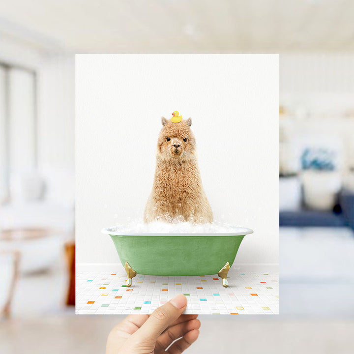 A hand holds up a framed image of a brown alpaca sitting in a green bathtub filled with bubbles, with a yellow rubber duck perched on its head.