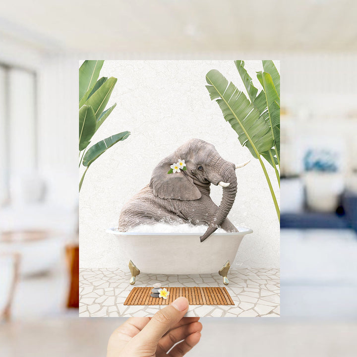A hand holds up a framed image depicting a gray elephant sitting in a white bathtub, surrounded by green leaves and flowers.
