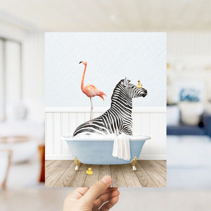 A hand holds up a framed image depicting a zebra sitting in a bathtub, with a pink flamingo standing on its back and a bird perched on its head.