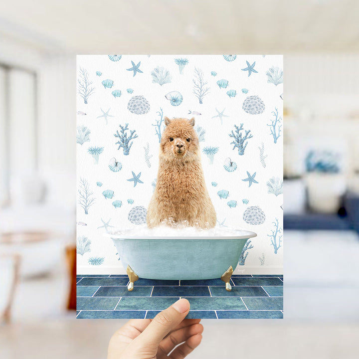 A hand is holding up a framed image of a brown alpaca sitting in a blue bathtub filled with bubbles, surrounded by a pattern of seashells, starfish, and other marine life.