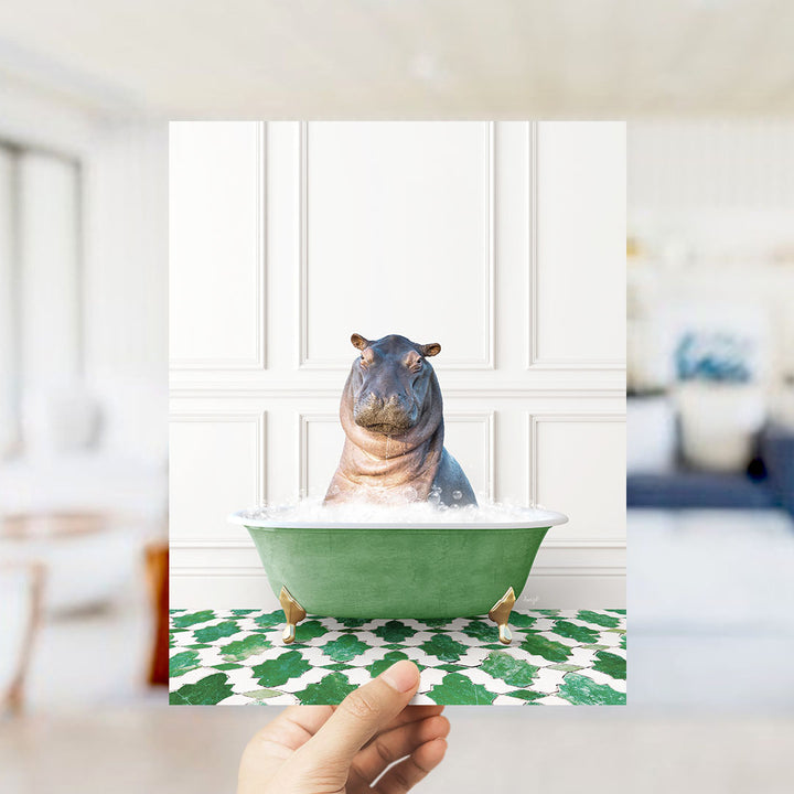 A hand holds up a framed image of a hippopotamus sitting in a green bathtub filled with bubbles, surrounded by a green and white patterned floor.