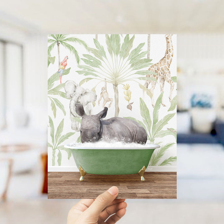 A hand holds up a framed print featuring a whimsical illustration of a rhinoceros bathing in a green bathtub, surrounded by tropical foliage and animals.