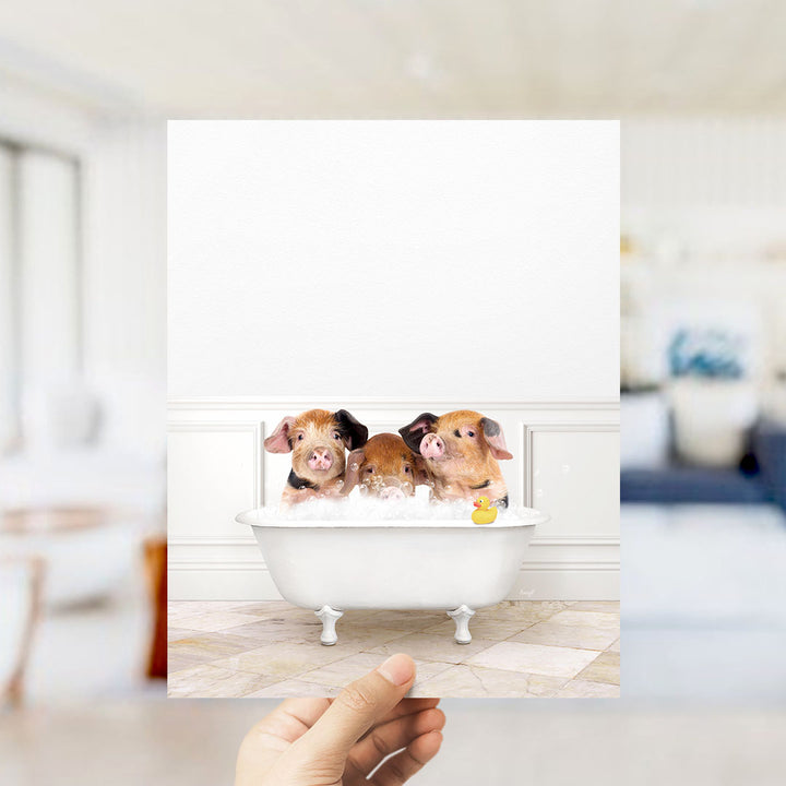A hand holds up a framed image of three adorable pigs sitting in a white bathtub, surrounded by bubbles and a rubber duck.