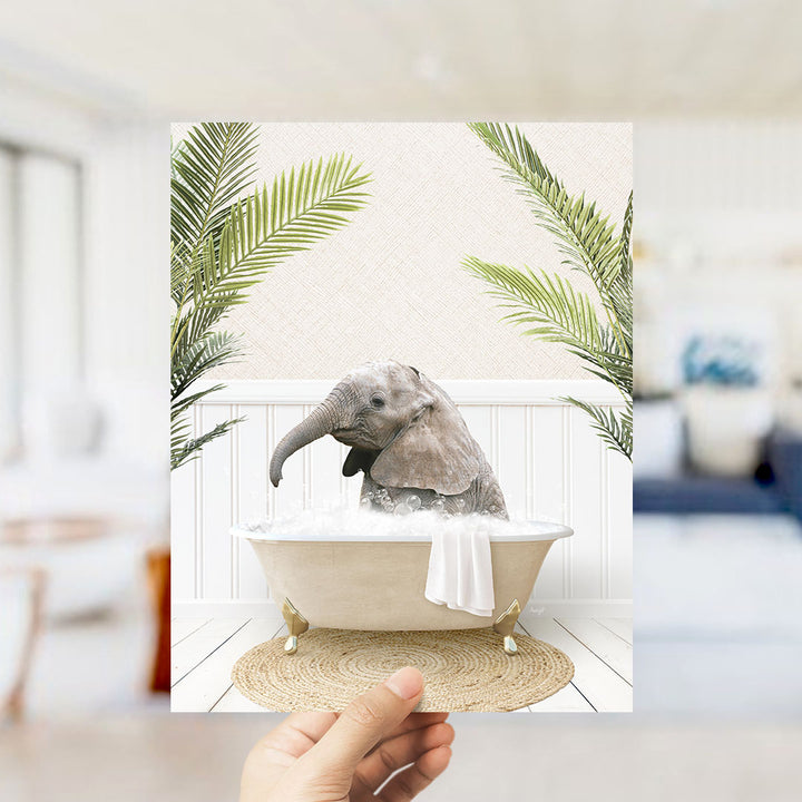 A hand holds up a framed image depicting a baby elephant sitting in a bathtub filled with water, surrounded by green palm leaves.
