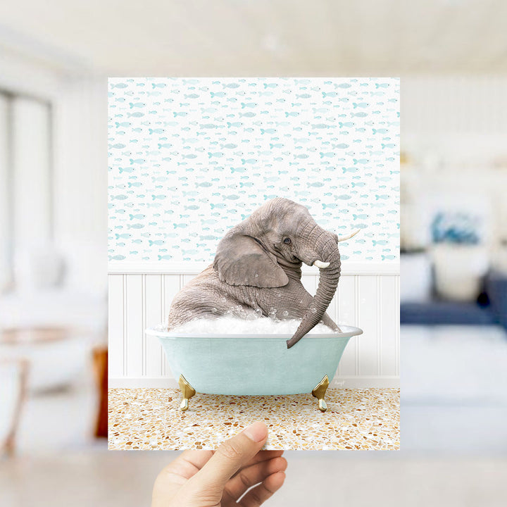 A hand holds up a framed image of a gray elephant sitting in a light blue bathtub filled with bubbles, surrounded by a pattern of small fish.