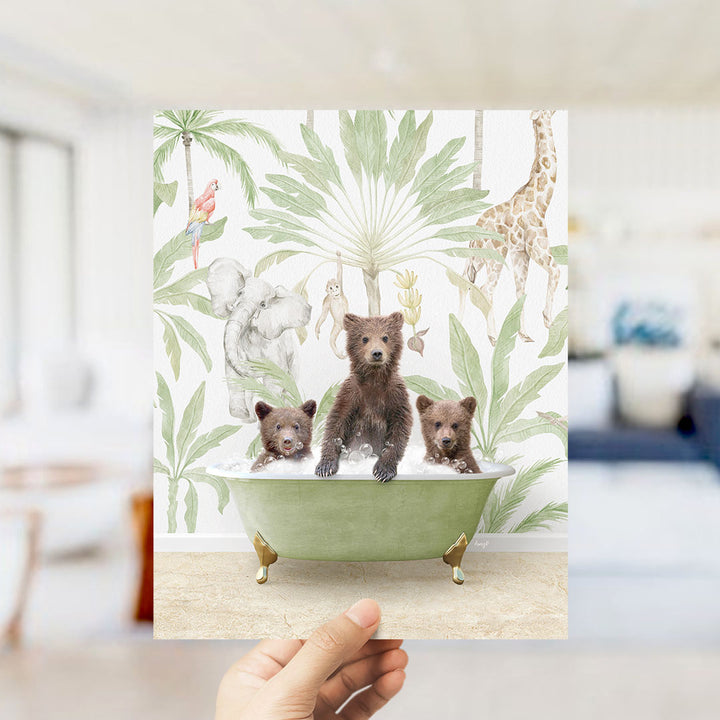 A hand holds up a framed print featuring a whimsical scene of three bears sitting in a bathtub, surrounded by tropical foliage and animals.