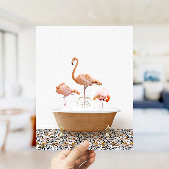 A hand holds up a framed image of three flamingos standing in a bathtub filled with water.