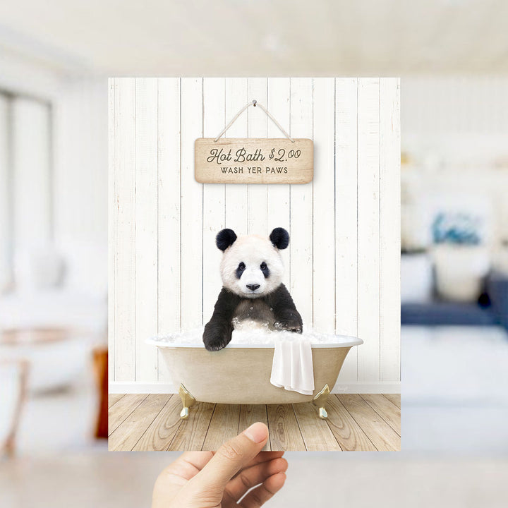 A hand holding a framed image of a panda bear sitting in a bathtub, with a sign above the bathtub that reads "Hot Bath $2.00 WASH YOUR PENGUINS".