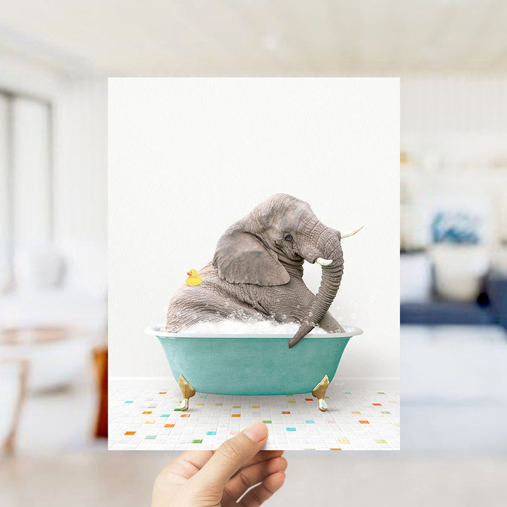 A hand holds up a framed image of a gray elephant sitting in a green bathtub, with a yellow rubber duck floating nearby.