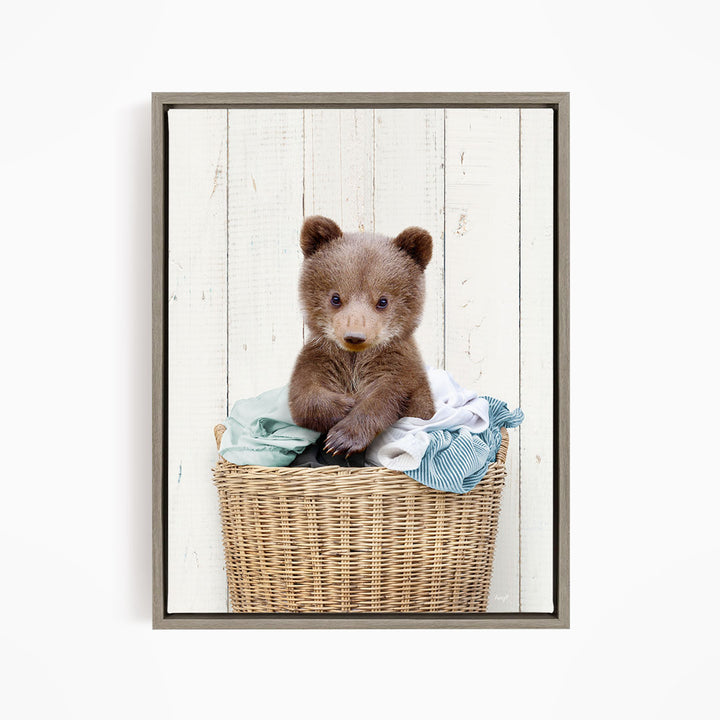 A young brown bear cub sitting in a wicker basket filled with clothes.