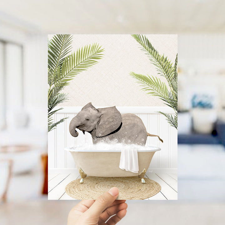 A hand holds up a framed image depicting a baby elephant sitting in a bathtub filled with water, surrounded by green palm leaves.