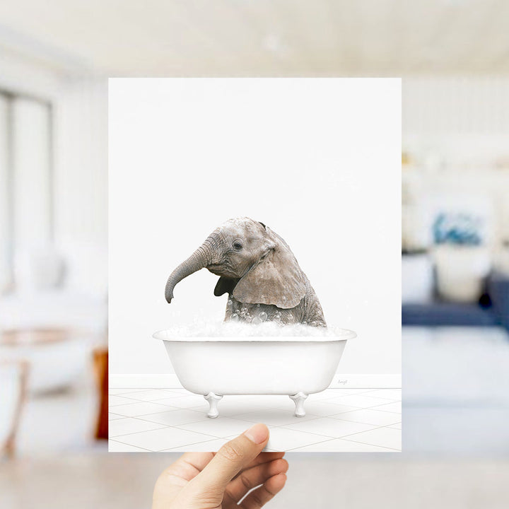 A hand holds up a framed image of a baby elephant sitting in a bathtub filled with water.