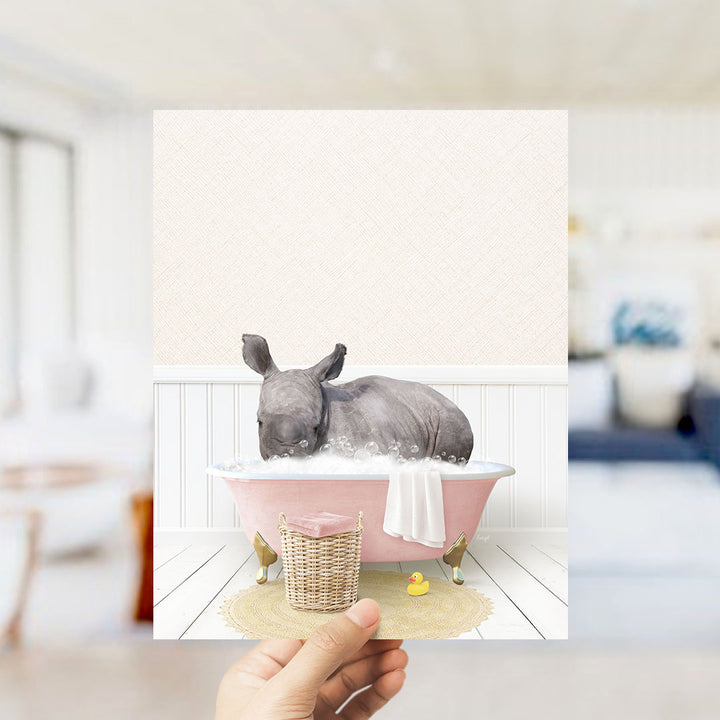 A rhinoceros is sitting in a pink bathtub filled with bubbles, surrounded by a yellow rubber duck and a white towel.