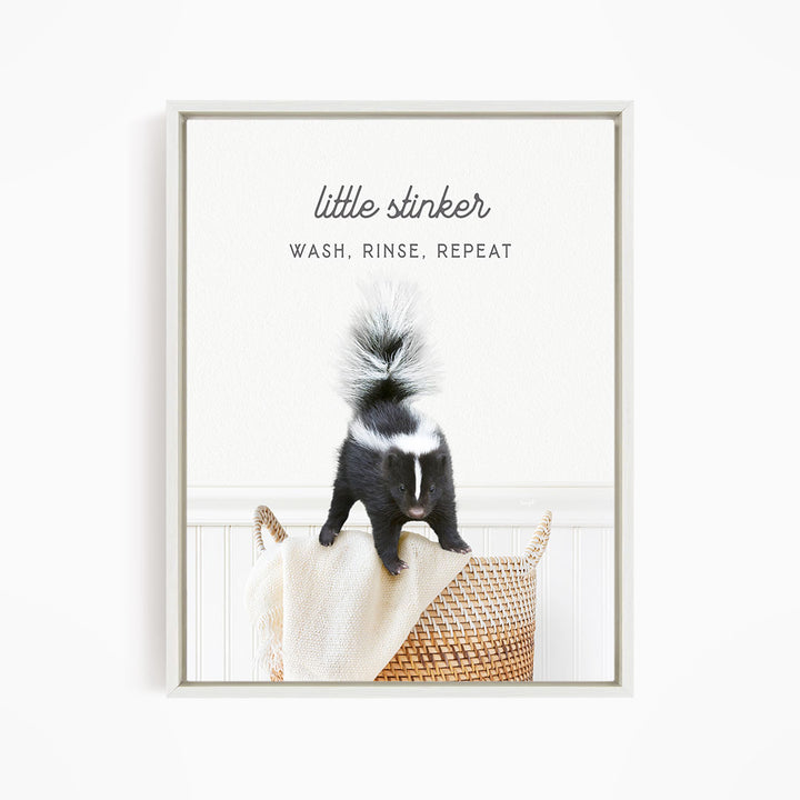 A black and white skunk standing on a woven basket, with the text "little stinker" and "WASH, RINSE, REPEAT" written above it.