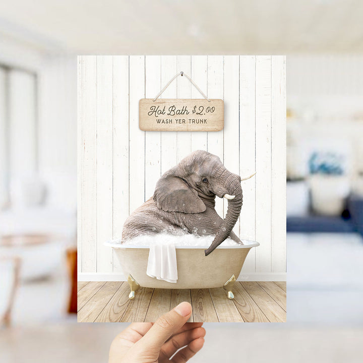 A hand holding a framed image of an elephant sitting in a bathtub, with a sign above it advertising a hot bath for $2.00.